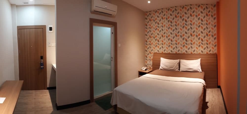 A cozy hotel room features a neatly made bed with white linens against an orange-patterned wall, a wooden desk, and a frosted glass door leading to a bathroom, ideal for travelers seeking comfort and simplicity.