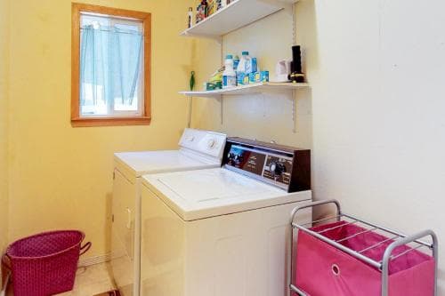 A cozy laundry room with yellow walls, a window with curtains, shelves stocked with supplies, and a washing machine paired with a dryer, ideal for travelers seeking self-sufficient accommodations.