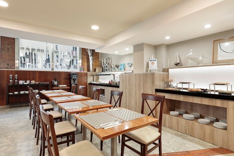 A cozy hotel dining area in Barcelona's Eixample district features neatly arranged wooden tables and chairs, a buffet counter with plates and decorative items, and a modern aesthetic ideal for travelers seeking a comfortable breakfast spot.