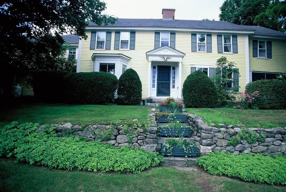 A charming yellow colonial-style house surrounded by lush greenery, stone walls, and vibrant flower beds creates a serene suburban retreat ideal for family vacations or weekend getaways.