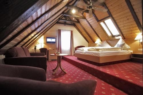 A cozy attic-style hotel room with wooden beams, red carpeting, comfortable seating, and a neatly made bed, ideal for travelers seeking a warm and inviting retreat.