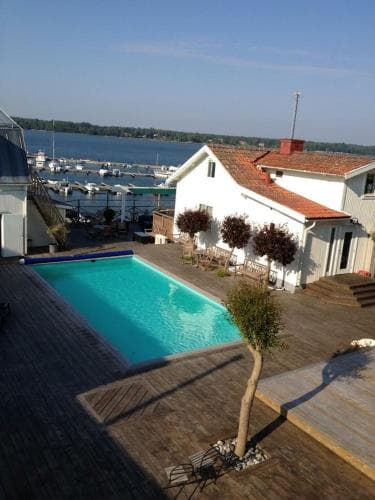 A serene lakeside villa features a vibrant blue pool, wooden deck, and charming red-roofed architecture, ideal for tranquil summer retreats near a marina.