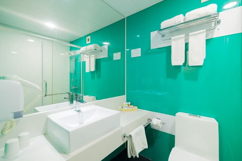 A modern bathroom at the Silvermine Beach Resort in Hong Kong features a vibrant teal wall, white fixtures, neatly arranged towels, and a hairdryer, ideal for travelers seeking comfort and cleanliness during their stay.