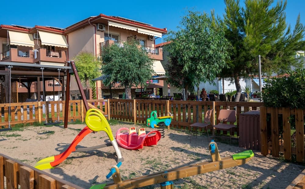 A vibrant playground with colorful toys and wooden fencing set against a backdrop of residential buildings and lush greenery, ideal for family-friendly vacation spots seeking a relaxing and playful atmosphere.
