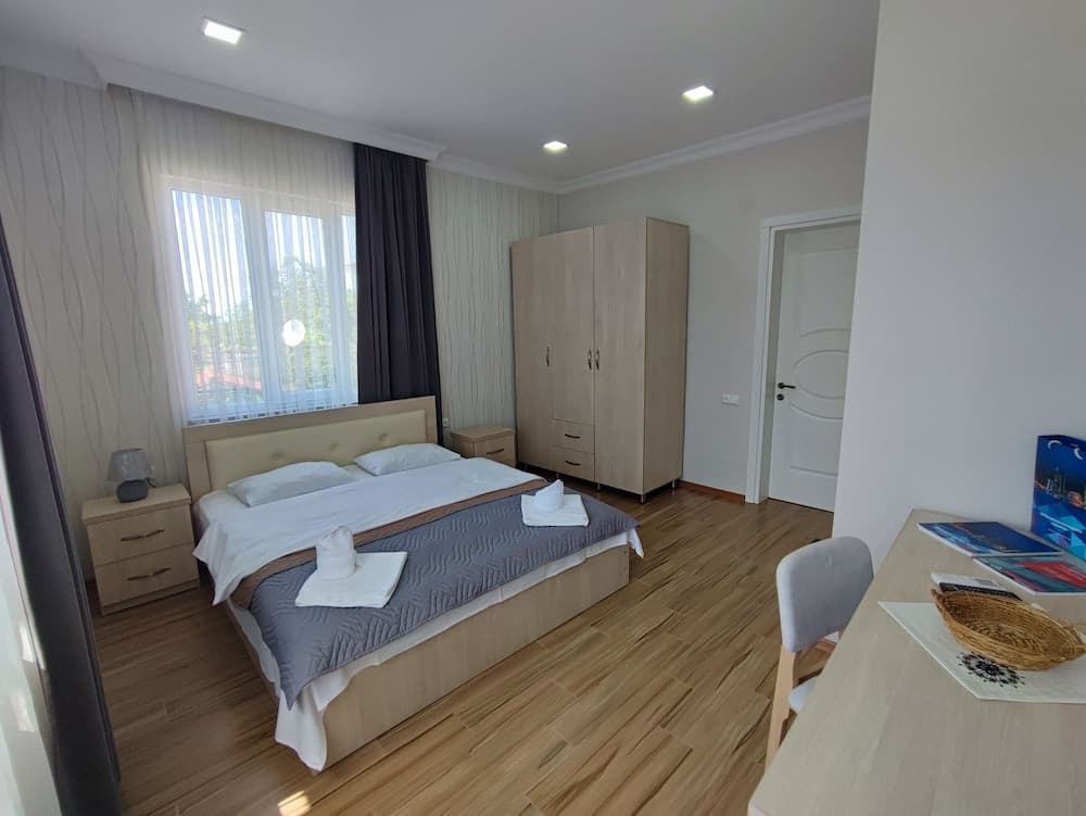 A cozy hotel room with wooden flooring, a neatly made bed adorned with folded towels, a large wardrobe, and a desk with travel brochures, ideal for travelers seeking comfort and convenience.