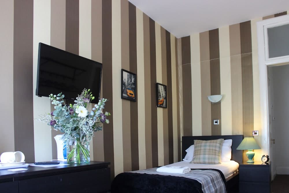 A cozy hotel room features striped walls, a neatly made bed with plaid pillows, a flat-screen TV, and a vase of fresh flowers, ideal for travelers seeking comfort and relaxation.