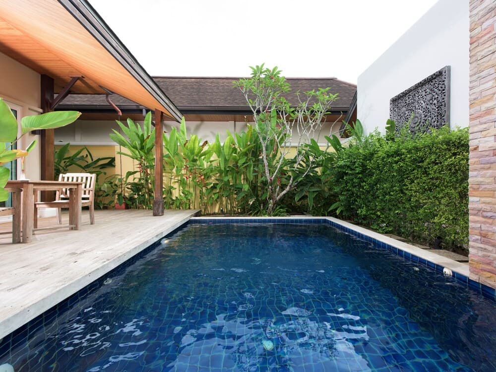 A serene tropical villa courtyard features a sparkling blue pool surrounded by lush greenery, wooden deck furniture, and intricate wall art, ideal for a relaxing vacation retreat.
