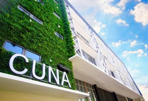 A vibrant urban hotel facade adorned with lush greenery and bold "CUNA" signage under a bright blue sky, ideal for eco-conscious travelers seeking modern accommodations.
