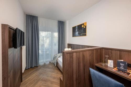 A cozy hotel room features a neatly made bed by a window with sheer curtains, a wooden desk with a chair, and modern decor, ideal for travelers seeking comfort and functionality.
