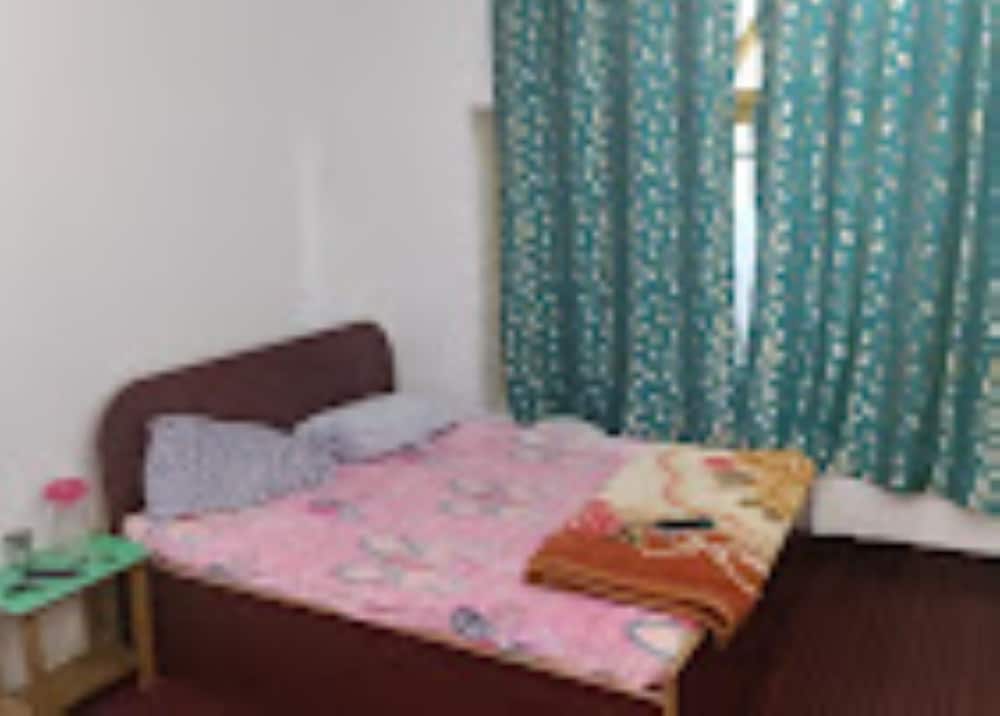 A cozy bedroom with a neatly made bed adorned in pink bedding, green patterned curtains, and a small side table creates a serene atmosphere ideal for travelers seeking a comfortable and budget-friendly accommodation.
