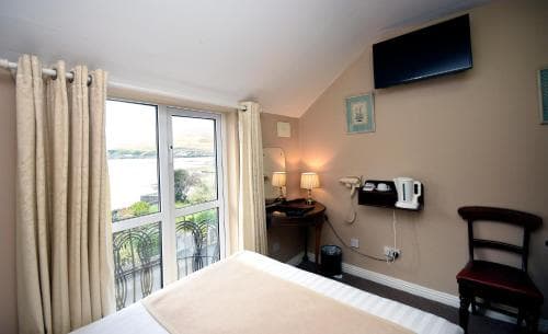 A cozy hotel room with a serene waterfront view through large windows, featuring a neatly made bed, a desk with lamps, and a mounted TV, ideal for travelers seeking relaxation and comfort.