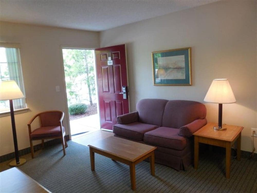 A cozy hotel room features a comfortable maroon sofa, wooden coffee table, and lamps, with a red door leading to a serene outdoor view, ideal for travelers seeking relaxation.
