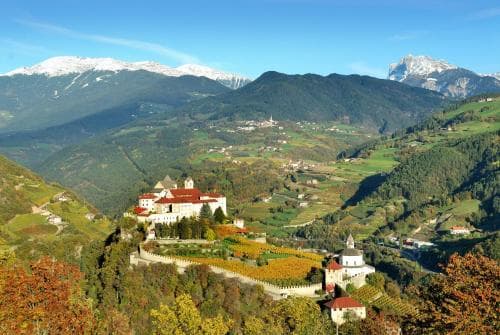 A picturesque medieval castle perched on a hillside overlooks a vibrant valley surrounded by snow-capped mountains, ideal for scenic photography and cultural exploration.