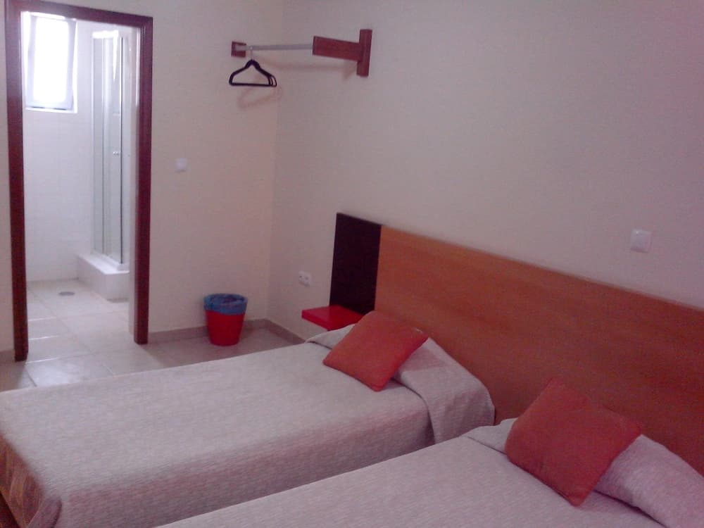 A cozy hotel room with two neatly made beds adorned with orange pillows, a red bucket near the entrance, and an adjacent bathroom, ideal for travelers seeking simple accommodations.