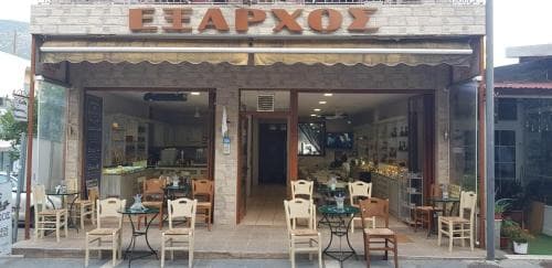 A cozy Greek café with outdoor seating invites travelers to enjoy its inviting atmosphere, featuring wooden chairs, tables, and a welcoming storefront perfect for a relaxing coffee break.