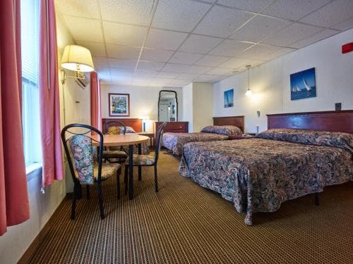 A cozy hotel room features two patterned beds, a small dining table by the window, warm lighting, and nautical artwork, ideal for travelers seeking comfort and convenience.