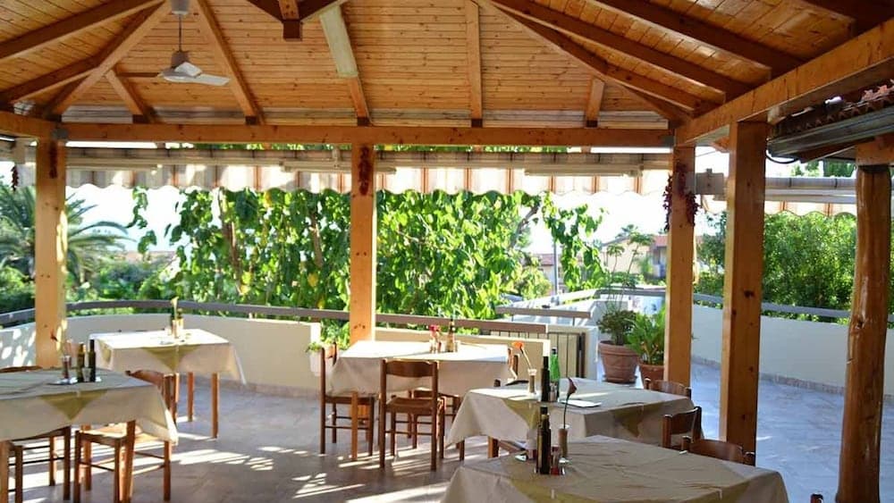 A serene outdoor dining area under a wooden pergola, surrounded by lush greenery, offers a tranquil spot for travelers to enjoy meals in a relaxed, sunlit setting.
