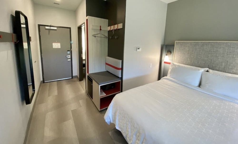 A modern hotel room features a cozy bed with white linens, a sleek seating area with red accents, and a full-length mirror, ideal for travelers seeking comfort and style during their stay.