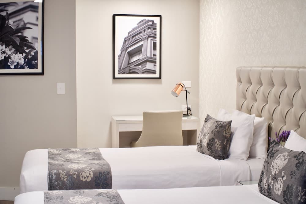 A cozy hotel room features a neatly made bed with floral-patterned accents, a modern desk setup with a stylish lamp, framed architectural art on the walls, and a serene ambiance ideal for travelers seeking comfort and elegance.