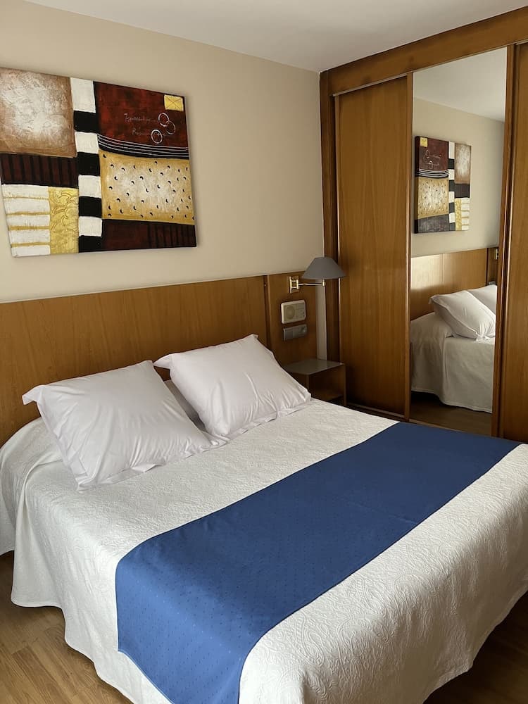 A cozy hotel room features a neatly made bed with white linens and a blue runner, complemented by wooden furnishings, a modern abstract painting, and a mirrored wardrobe, ideal for travelers seeking comfort and style.