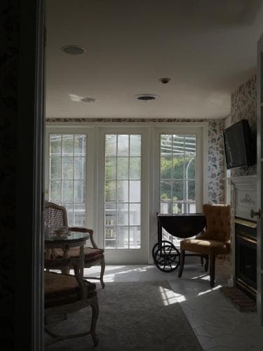 A cozy indoor lounge area features a wheelchair, vintage chairs, and a large window with French doors leading to a bright balcony, ideal for travelers seeking accessible accommodations in a serene setting.