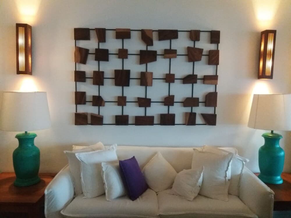 A cozy living room features a white sofa adorned with plush pillows, flanked by teal lamps on wooden tables, under a geometric wooden wall art illuminated by warm sconces, ideal for travelers seeking a serene and stylish accommodation.