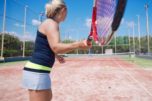 A tennis player practices her swing on an outdoor clay court under a sunny sky, ideal for sports enthusiasts seeking active vacation activities.