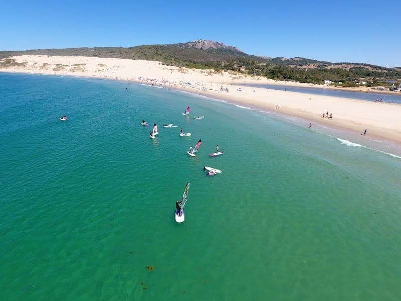 A vibrant beach scene at Playa de Valdevaqueros in Cádiz, Spain, showcases windsurfers and wingfoilers enjoying the turquoise waters under a clear blue sky, ideal for adventure seekers looking to explore water sports destinations.