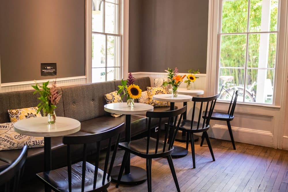 A cozy café corner with wooden floors, cushioned benches adorned with floral pillows, small round tables topped with vibrant sunflowers, and large windows letting in natural light, perfect for a relaxing coffee break or a quiet work session.
