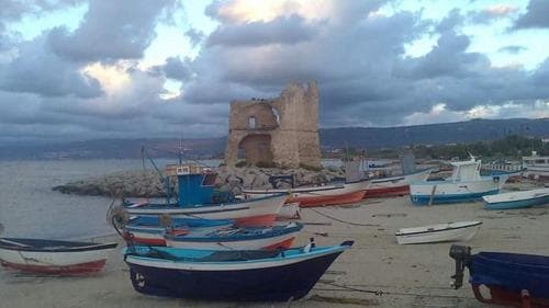 A serene coastal scene features colorful fishing boats docked on sandy shores under a dramatic sky, with a historic stone tower in the background, ideal for travelers seeking picturesque seaside escapes.