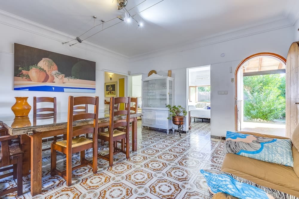 A cozy living and dining area with patterned tile floors, wooden furniture, vibrant artwork, and an inviting view of lush greenery through arched windows, perfect for travelers seeking a charming and comfortable vacation rental.