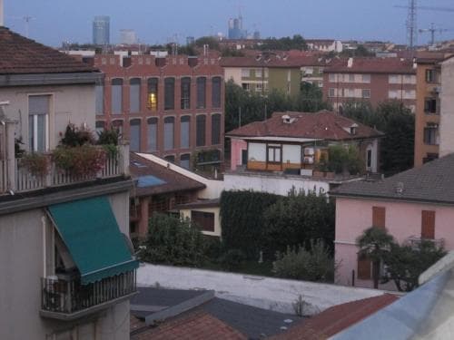 A serene rooftop view of residential buildings with terracotta roofs, lush greenery, and distant skyscrapers under a clear sky, ideal for urban exploration and photography enthusiasts.