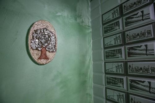 A serene indoor gallery corner features a textured tree sculpture on a green wall beside a tiled display of black-and-white cityscape prints, ideal for art enthusiasts exploring urban-themed exhibits.