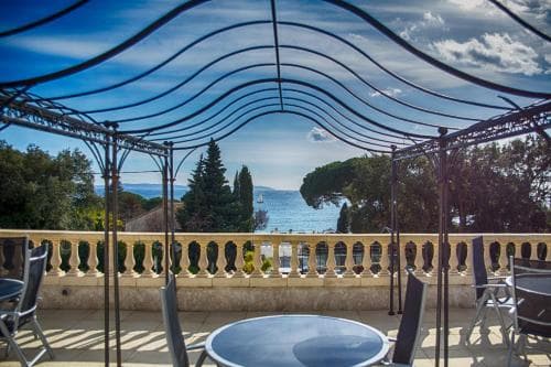 A serene coastal terrace with a metal pergola offers a picturesque view of the sea, ideal for relaxing getaways amidst lush greenery and clear skies.