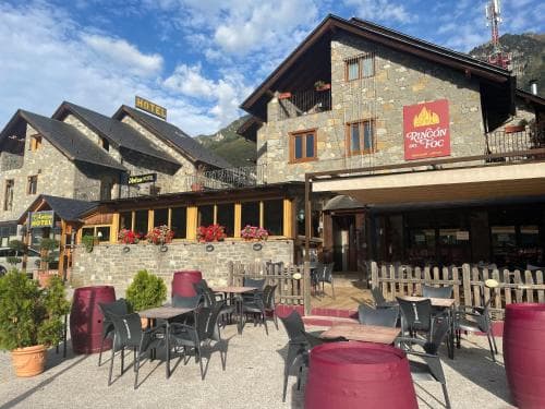 A charming mountain village scene features rustic stone buildings with outdoor seating, vibrant red flowers, and a cozy atmosphere ideal for travelers seeking relaxation in a picturesque alpine setting.