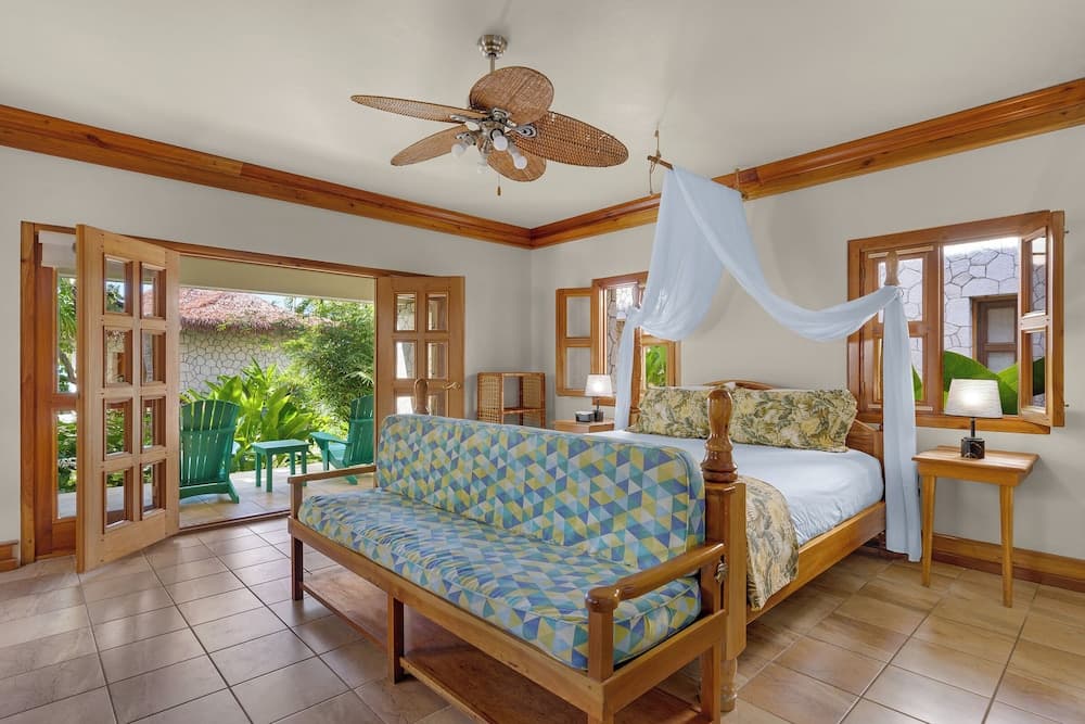 A cozy tropical bedroom with wooden furnishings, a patterned sofa, a canopy bed, and open doors leading to a lush garden, ideal for travelers seeking a serene and inviting retreat.
