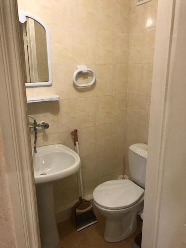 A compact bathroom with beige tiled walls, a sink, toilet, mirror, and cleaning tools creates a functional and practical space ideal for travelers seeking efficient accommodations.