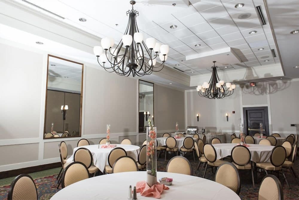 A spacious banquet hall adorned with elegant chandeliers, round tables set with white linens and floral centerpieces, and large mirrors reflecting the serene ambiance, ideal for hosting weddings or corporate events.
