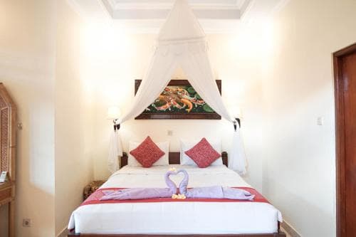 A cozy hotel room features a neatly made bed adorned with decorative pillows and a swan-shaped towel arrangement under a white canopy, set against warm walls with a framed artwork, ideal for travelers seeking comfort and charm.
