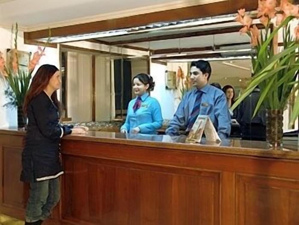 A warmly lit hotel reception area features attentive staff in blue uniforms assisting guests at a polished wooden counter adorned with vibrant floral arrangements, ideal for travelers seeking a welcoming check-in experience.