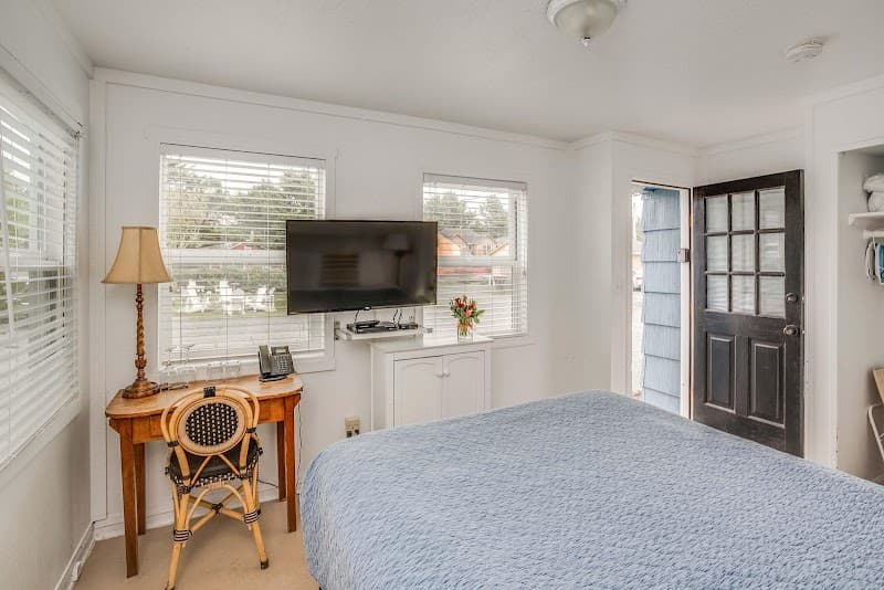 A cozy hotel room in Cannon Beach features a neatly made bed, a wooden desk with a chair, a flat-screen TV, and large windows offering a serene view, ideal for travelers seeking relaxation and comfort.