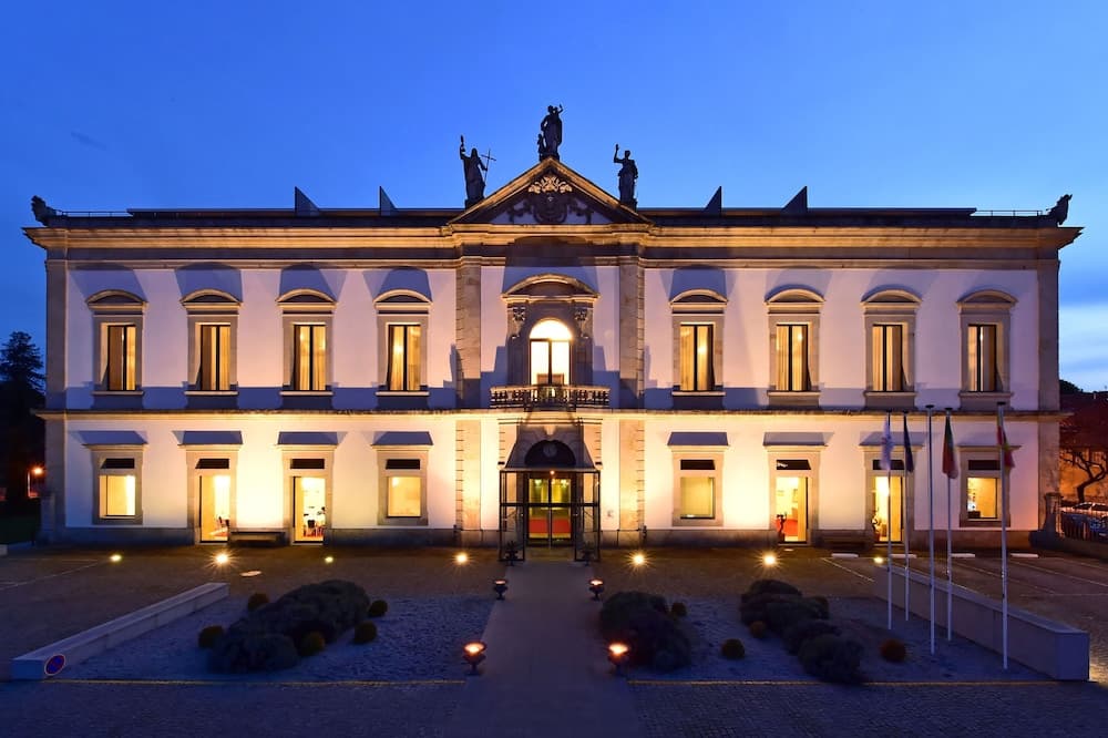 A grand, illuminated neoclassical building with statues and flags stands majestically at dusk, offering a serene and elegant backdrop for luxury travel experiences.