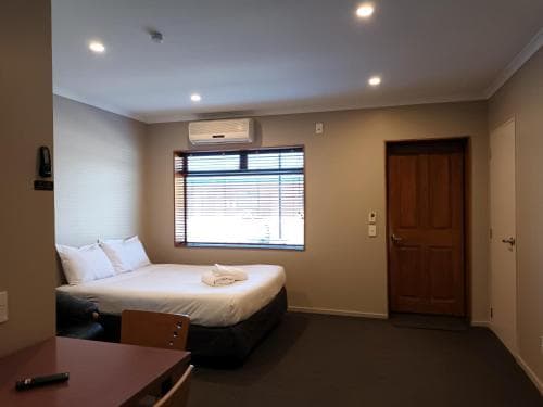 A cozy hotel room features a neatly made bed by a window with blinds, a wooden door, and simple furnishings, creating a calm and inviting atmosphere ideal for travelers seeking comfort and relaxation.