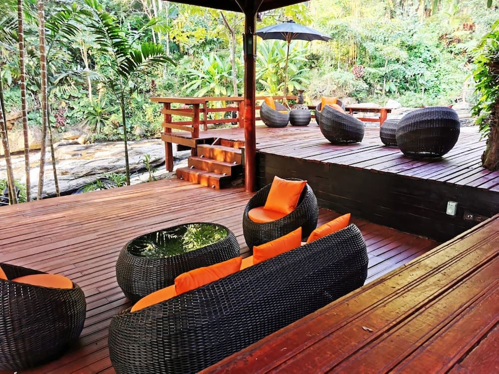 A serene tropical retreat features a wooden deck adorned with cozy wicker furniture and vibrant orange cushions, surrounded by lush greenery, ideal for relaxing vacations or nature escapes.