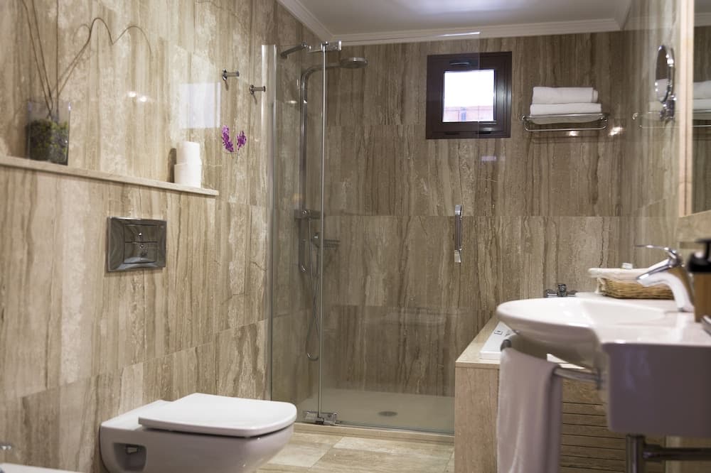 A modern bathroom with beige marble tiles features a sleek walk-in shower, a wall-mounted TV, and a minimalist sink setup, ideal for travelers seeking luxury and relaxation.