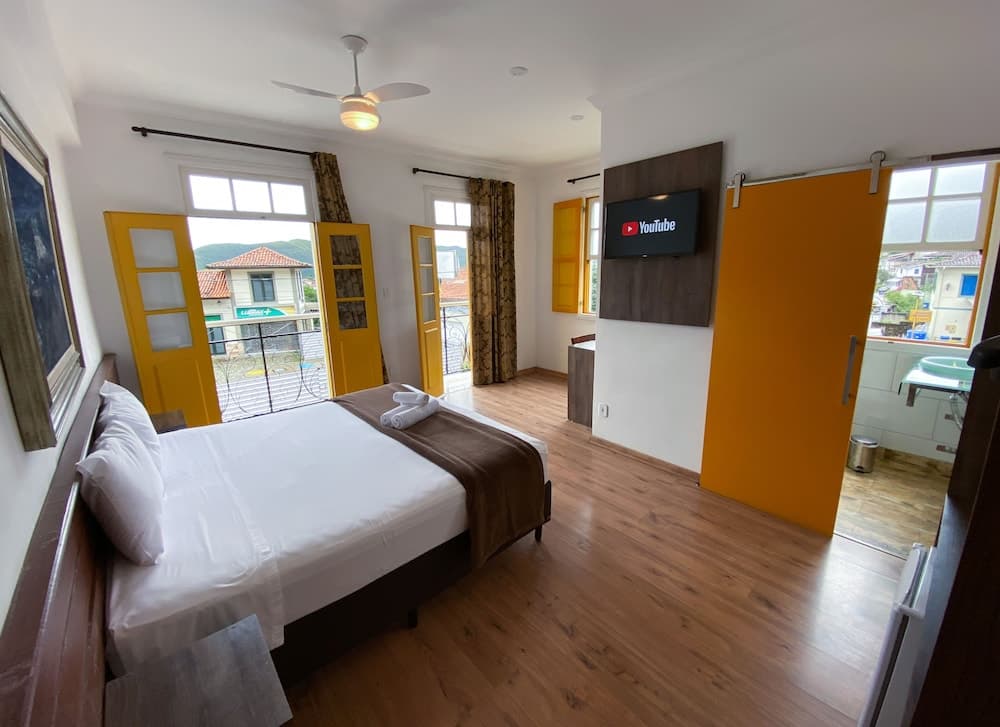 A cozy hotel room with wooden floors, vibrant yellow accents, and a view of rooftops through large windows offers a comfortable stay for travelers seeking relaxation and modern amenities.