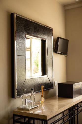 A cozy hotel bathroom features a stylish black-framed mirror reflecting natural light, a sleek countertop with toiletries, and a mounted TV, ideal for travelers seeking modern amenities in a serene setting.