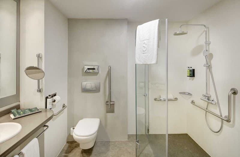 A modern, accessible hotel bathroom in Barcelona's Eixample district features sleek fixtures, a glass-enclosed shower, and thoughtful amenities, ideal for travelers seeking comfort and convenience.