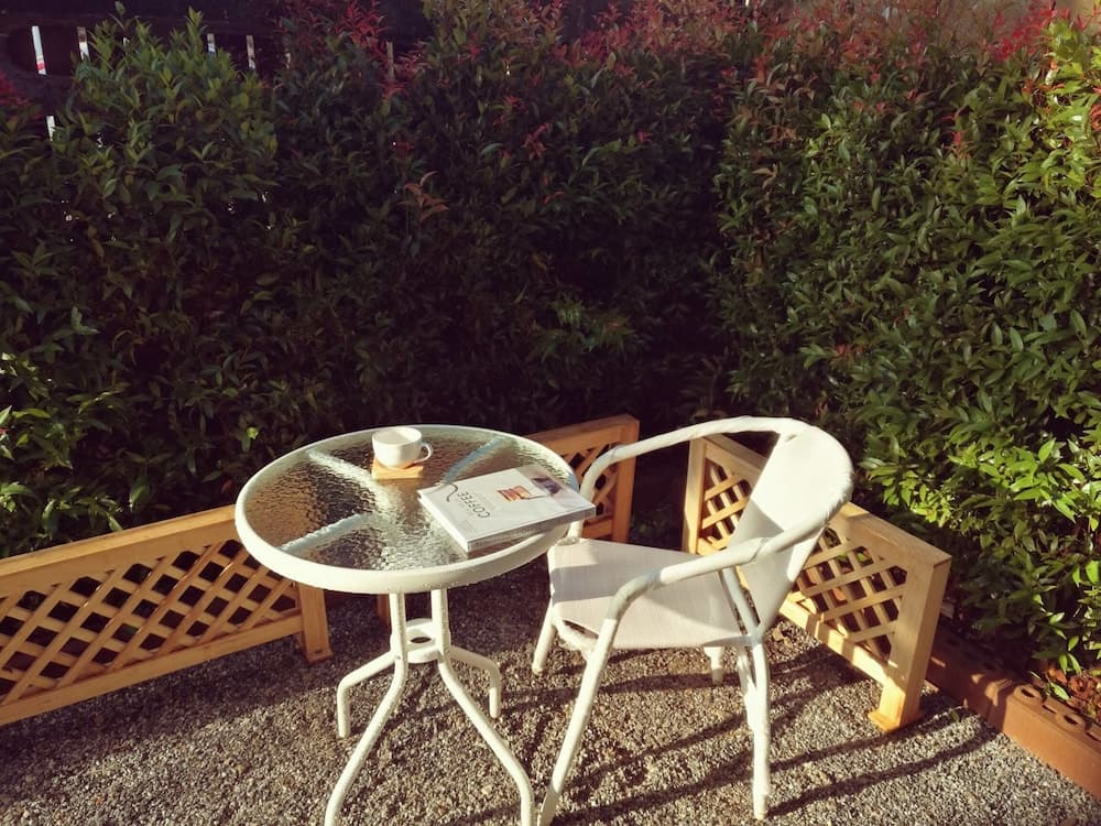 A serene outdoor patio features a white table set with a coffee cup and magazine, surrounded by lush greenery, ideal for a peaceful morning retreat or a cozy vacation spot.
