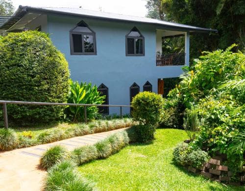A serene tropical garden surrounds a charming light blue house with lush greenery, ideal for eco-friendly travelers seeking a peaceful retreat.